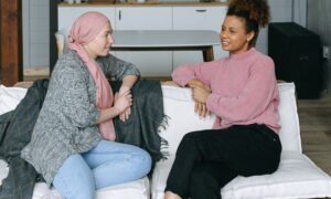 Two women talk on a couch, one is wearing a head covering.